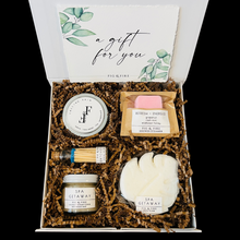 Load image into Gallery viewer, The petite self-care gift box is pictured. It contains a silver candle tin, matches, a shower steamer, a bath bomb, and a small sugar scrub.