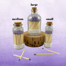 Load image into Gallery viewer, Image of the three sizes of corked glass bottles containing lavender purple matchsticks for The Breasties. There are a few loose purple matchsticks outside of the bottles. One of the large match bottles is on a cork. Background image has different shades of lavender purple.