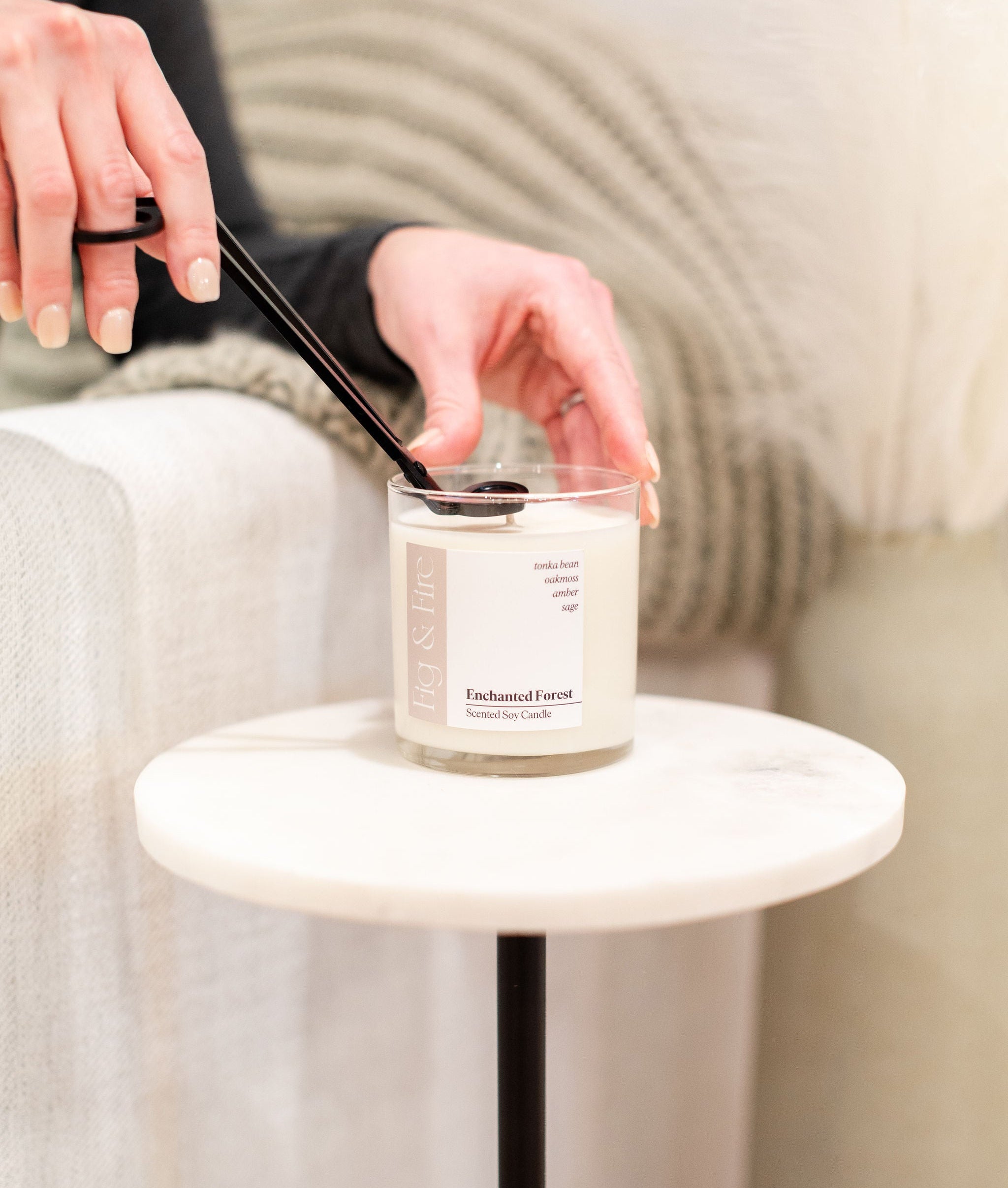 Hands using wick trimmers to trim a candle labeled 'Enchanted Forest' on a small round table.