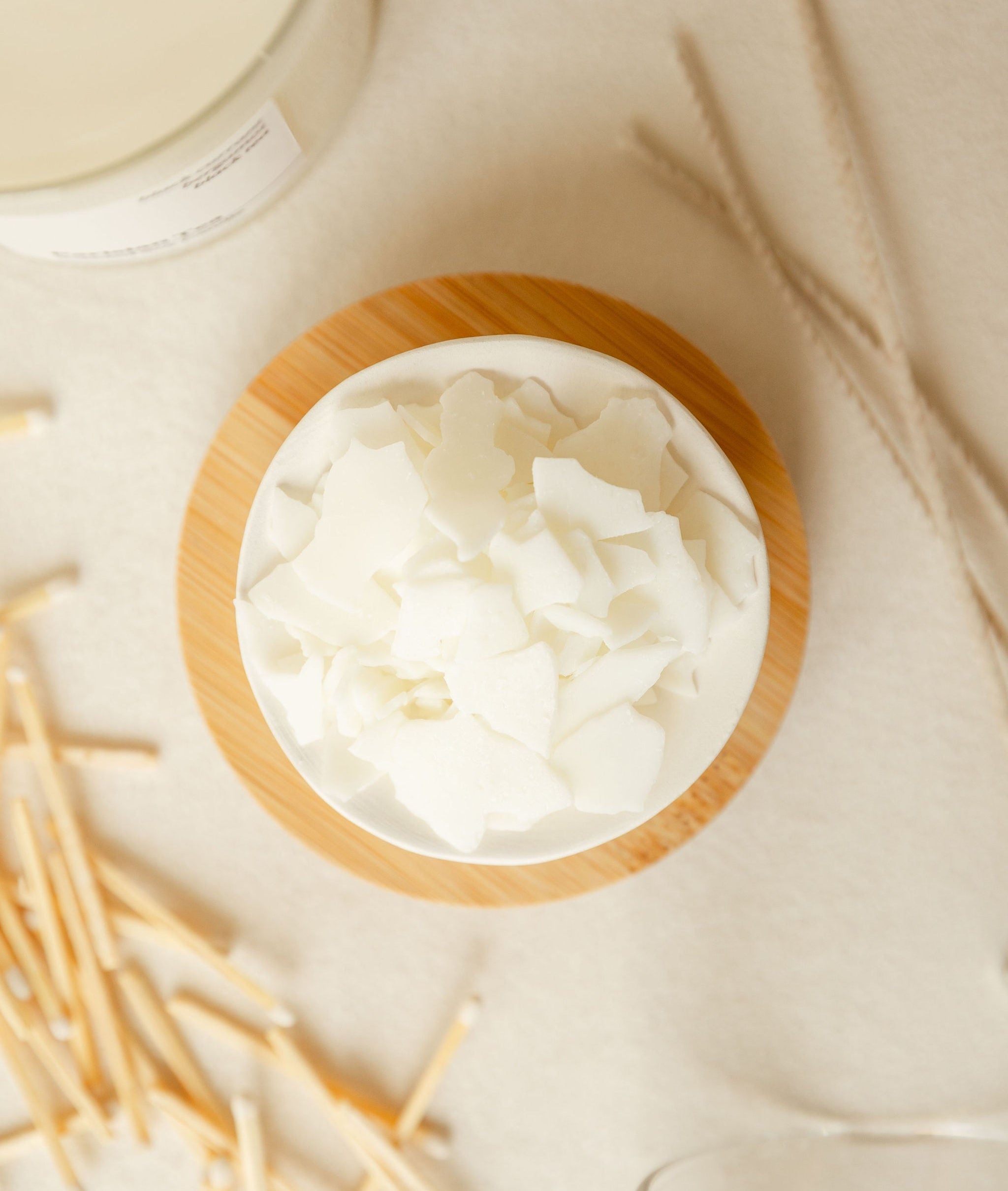 Wax pieces on a white dish, on top of a bamboo wooden lid, next to matchsticks, wicks, and glass candles