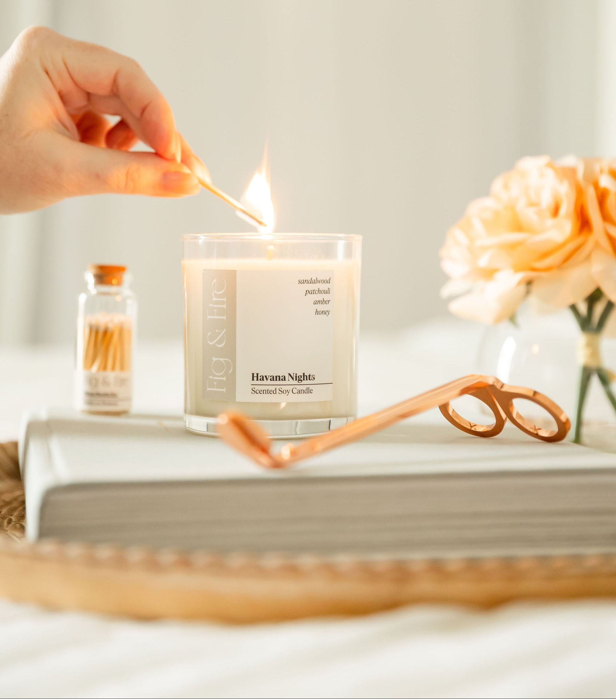 Hand lighting a candle labeled 'Havana Nights' with a book, matches, wick trimmers, and flowers in the background.