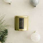 Winter Woods gold candle tin with a black label on a beige surface with Christmas / holiday decorations