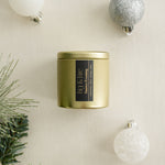 Snowy Evening gold candle tin with a black label on a beige surface with Christmas / holiday decorations
