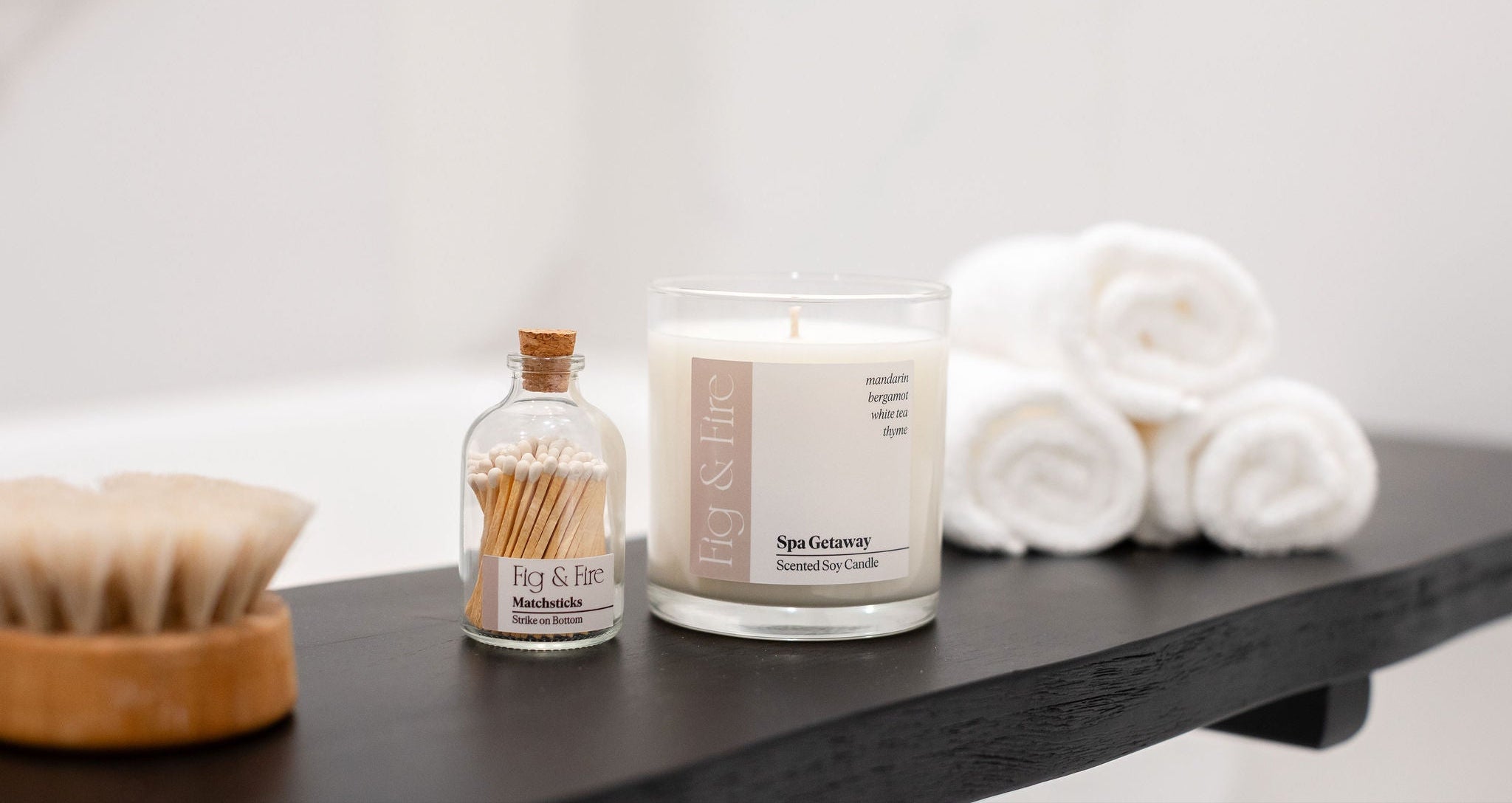 Candle, bottle of matches, and folded towels on a wooden surface with a neutral background