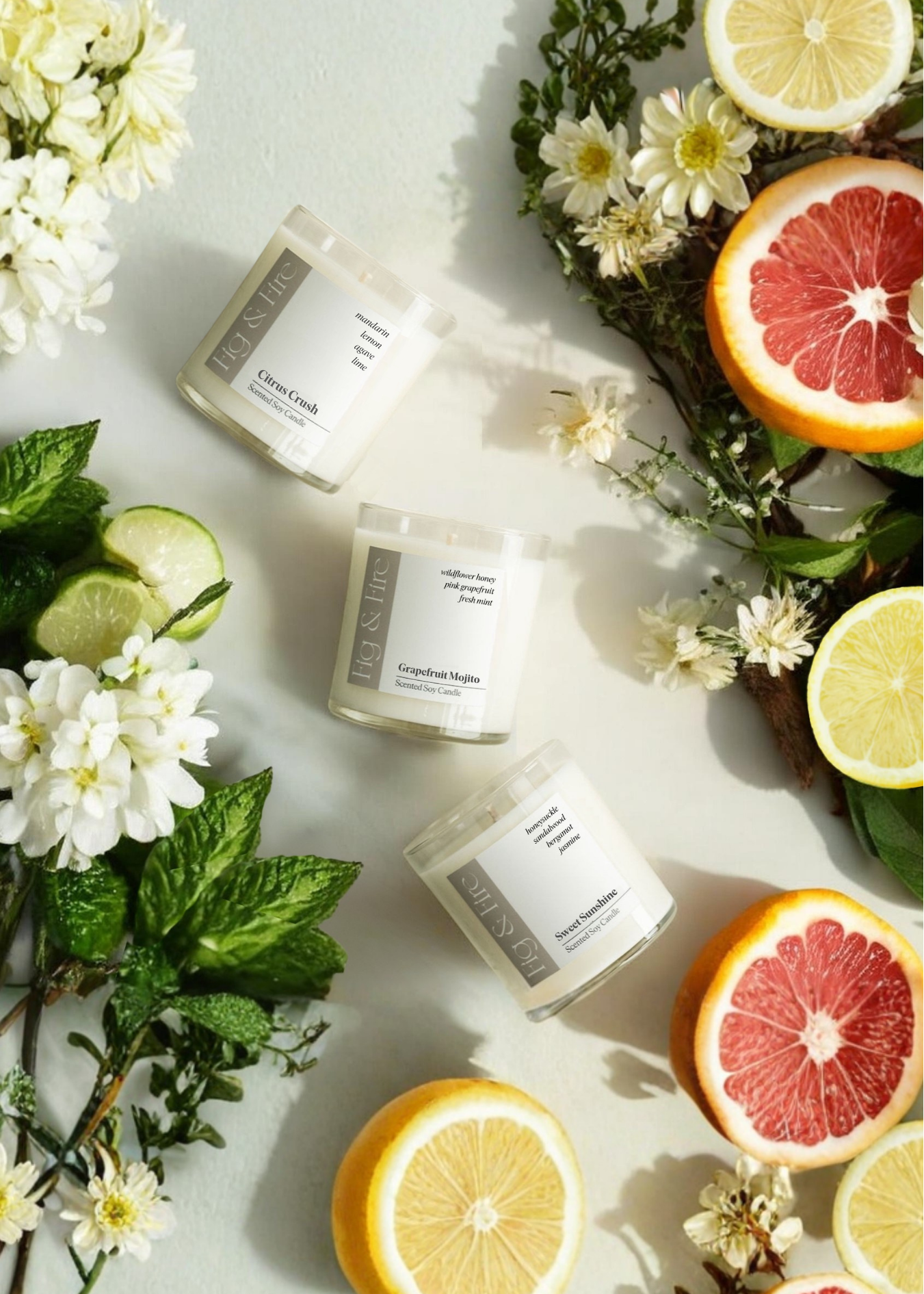 The entire spring collection is featured - Citrus Crush, Grapefruit Mojito, Sweet Sunshine - the vessels are clear glass tumblers - all-natural, vegan, non-toxic soy candles made with essential oils. the background has florals and citrus fruits, as well as greenery