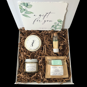 The petite self-care gift box is pictured. It contains a silver candle tin, matches, a shower steamer, and a small sugar scrub.