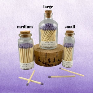 Image of the three sizes of corked glass bottles containing lavender purple matchsticks for The Breasties. There are a few loose purple matchsticks outside of the bottles. One of the large match bottles is on a cork. Background image has different shades of lavender purple.
